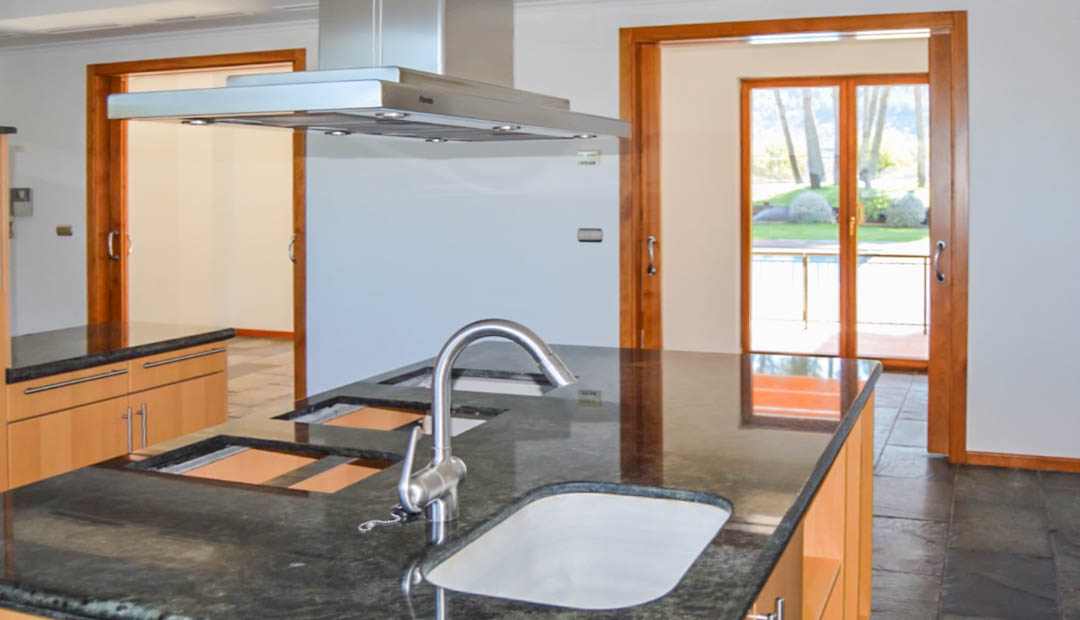 Cocina moderna con isla central de granito oscuro, campana extractora de acero y suelos de piedra natural. Incluye acceso directo al exterior mediante puertas correderas de madera.