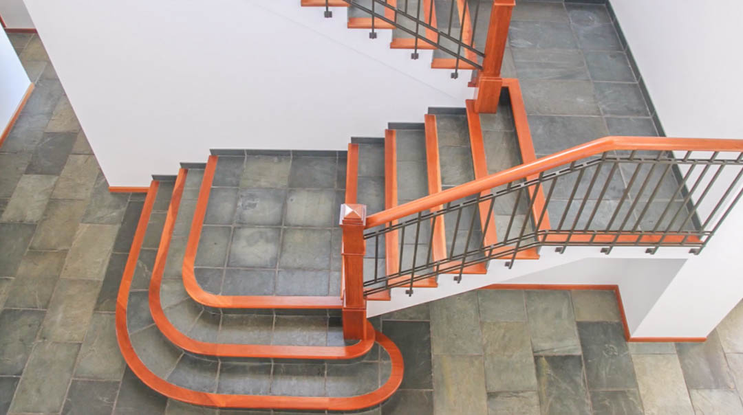 Escalera principal con peldaños de piedra natural y bordes de madera noble. Destaca la barandilla de hierro forjado con pasamanos de madera sobre suelos de baldosa oscura.
