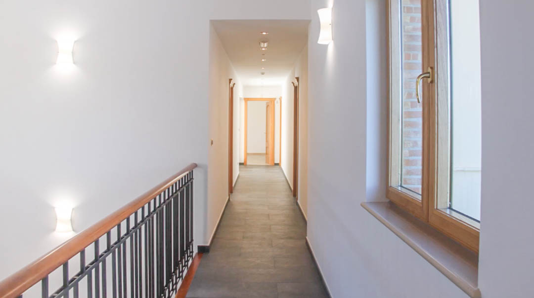 Pasillo luminoso con suelos de piedra gris, carpintería de madera de alta calidad y una barandilla de forja y madera que asoma a la escalera principal.