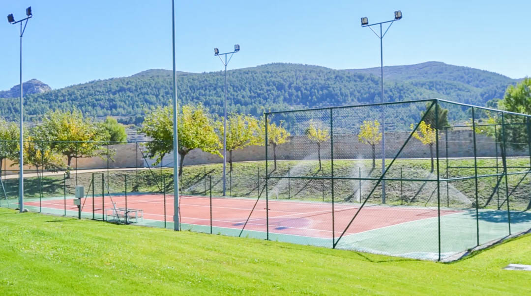 Pista de tenis vallada con superficie roja y verde, equipada con potentes torres de iluminación y rodeada de jardines con vistas a las montañas.