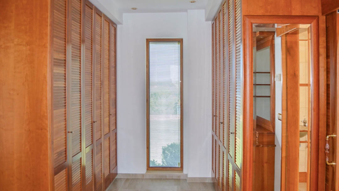 Vestidor con armarios empotrados de madera de alta calidad y puertas de persiana, iluminado por luz natural a través de un ventanal vertical y focos empotrados.