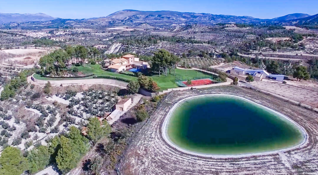 Vista aérea de la finca con la villa principal, piscina, pista de tenis y un gran embalse privado, rodeados de extensos campos de cultivo y montañas.