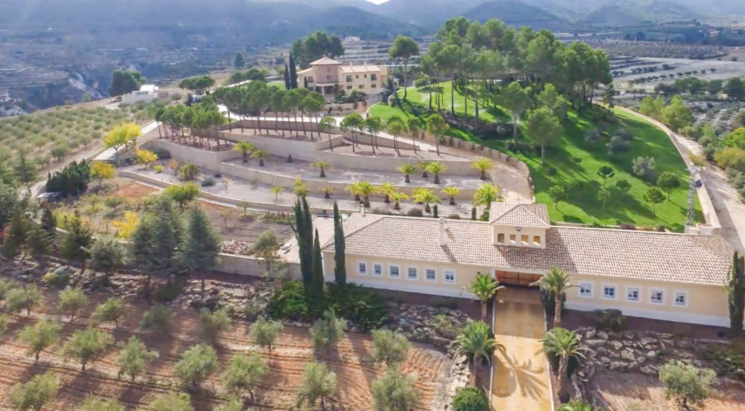 Vista aérea de la finca que muestra el almacén principal, jardines aterrazados con palmeras y la villa al fondo, rodeada de extensos olivares y montañas.