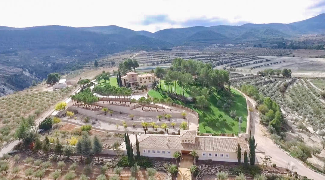 Vista aérea de la finca que muestra el chalet principal, la casa de entrada y el paisajismo aterrazado rodeado de extensos campos de cultivo y montañas.