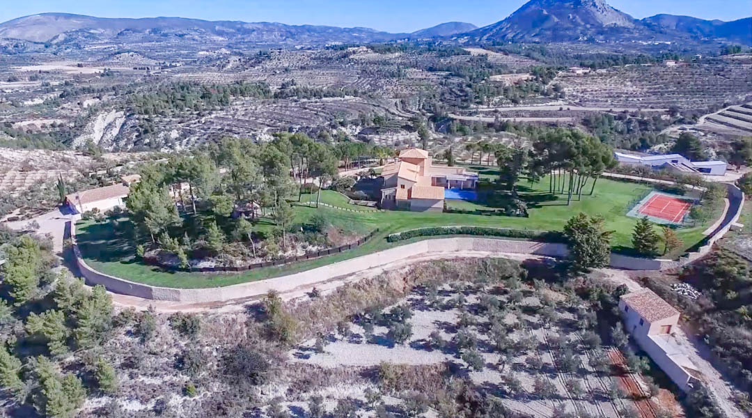 Vista aérea de la finca que muestra la villa principal, la piscina y la pista de tenis privada, rodeadas por extensos jardines y campos de cultivo en terrazas.