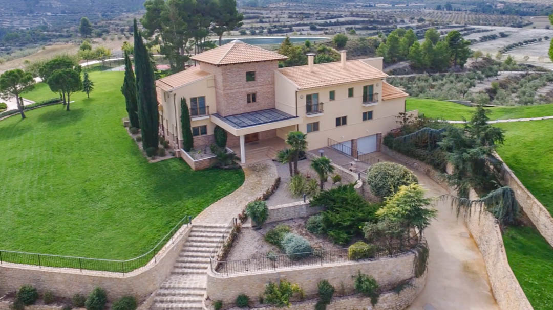 Vista aérea de la villa principal con fachada de piedra y estuco, rodeada de jardines cuidados, muros de mampostería y extensos campos de cultivo en un entorno montañoso.