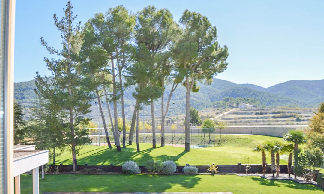 Vista desde la planta superior hacia el jardín con césped, pinos altos, palmeras y una pista de tenis al fondo, rodeada de un paisaje montañoso.