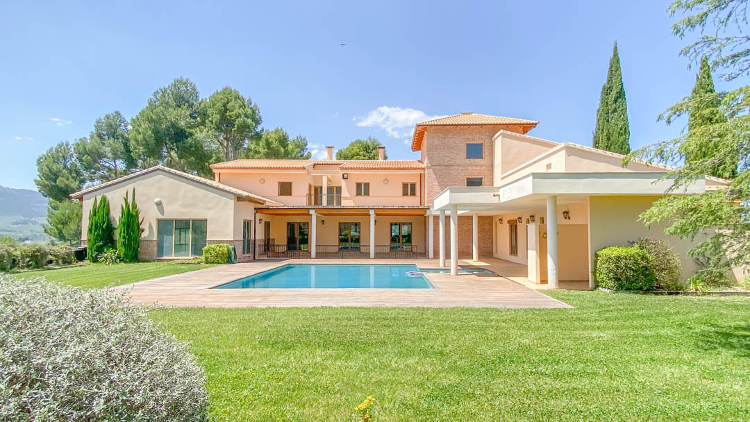 Vista exterior de la villa principal con piscina rectangular, rodeada de amplias zonas verdes y porches con columnas bajo un cielo despejado.