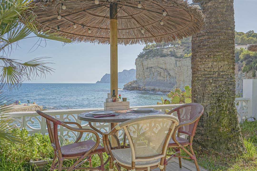 Comedor exterior bajo sombrilla de brezo con vistas frontales al mar y los acantilados. Incluye mobiliario de estilo bistró junto a una palmera y barandilla clásica.