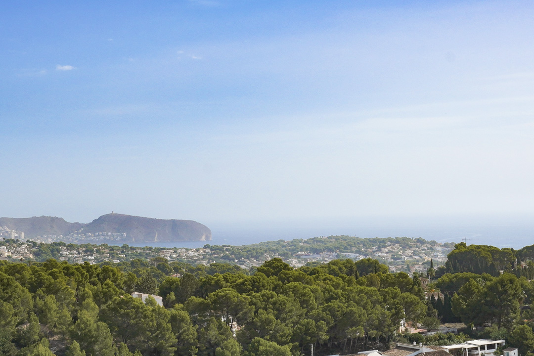 Vista panorámica elevada hacia el Cap D'or en Moraira, mostrando la densa vegetación de pinos mediterráneos y la costa bajo un cielo despejado.