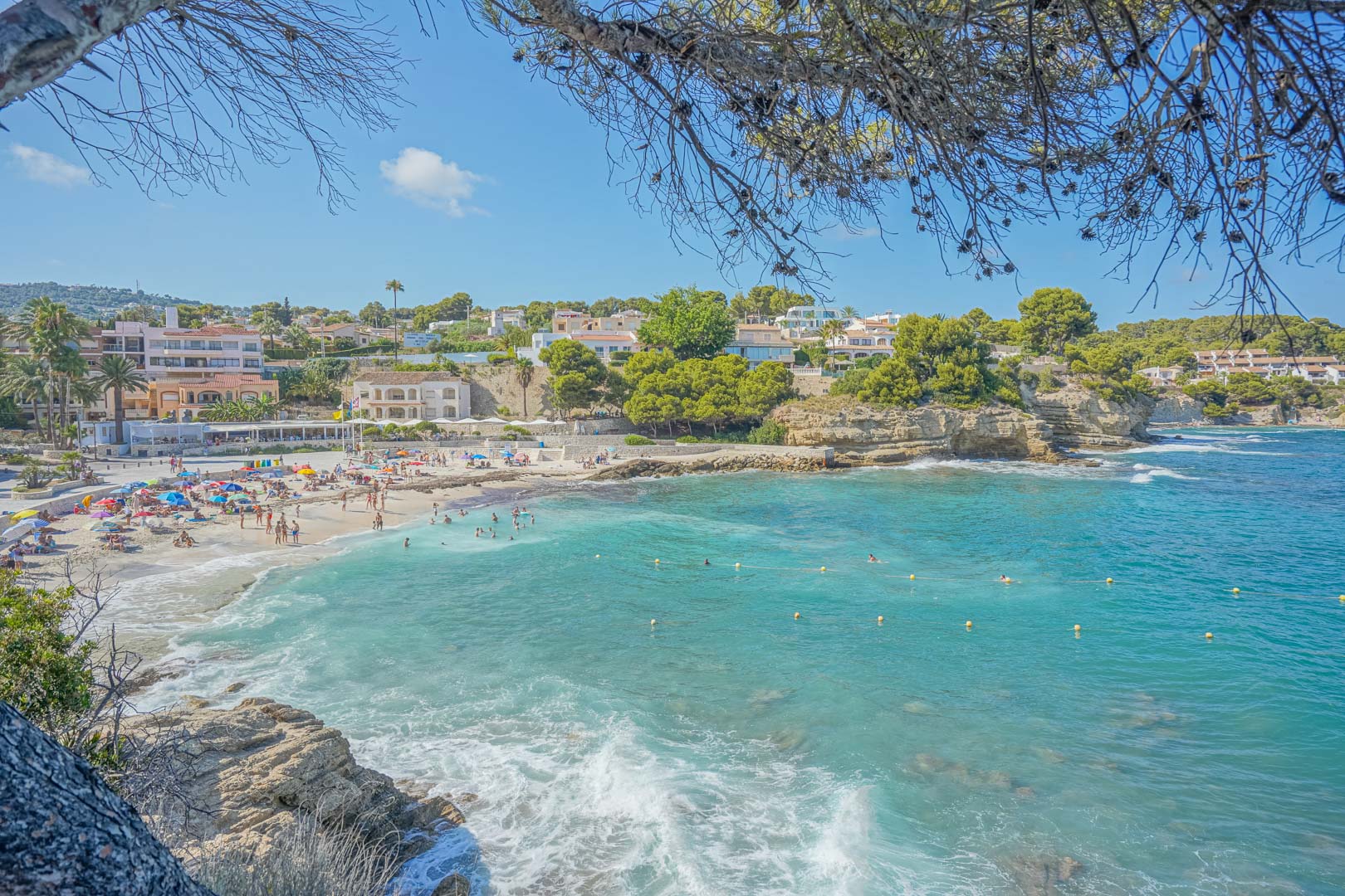Vista panorámica de una cala de aguas cristalinas en Benissa, rodeada de acantilados rocosos, pinos mediterráneos y edificaciones costeras bajo un cielo despejado.