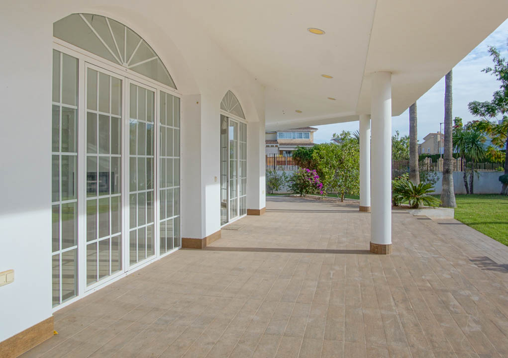 Amplia terraza cubierta con suelos cerámicos, columnas blancas y grandes ventanales de arco que conectan el interior con el jardín de palmeras.