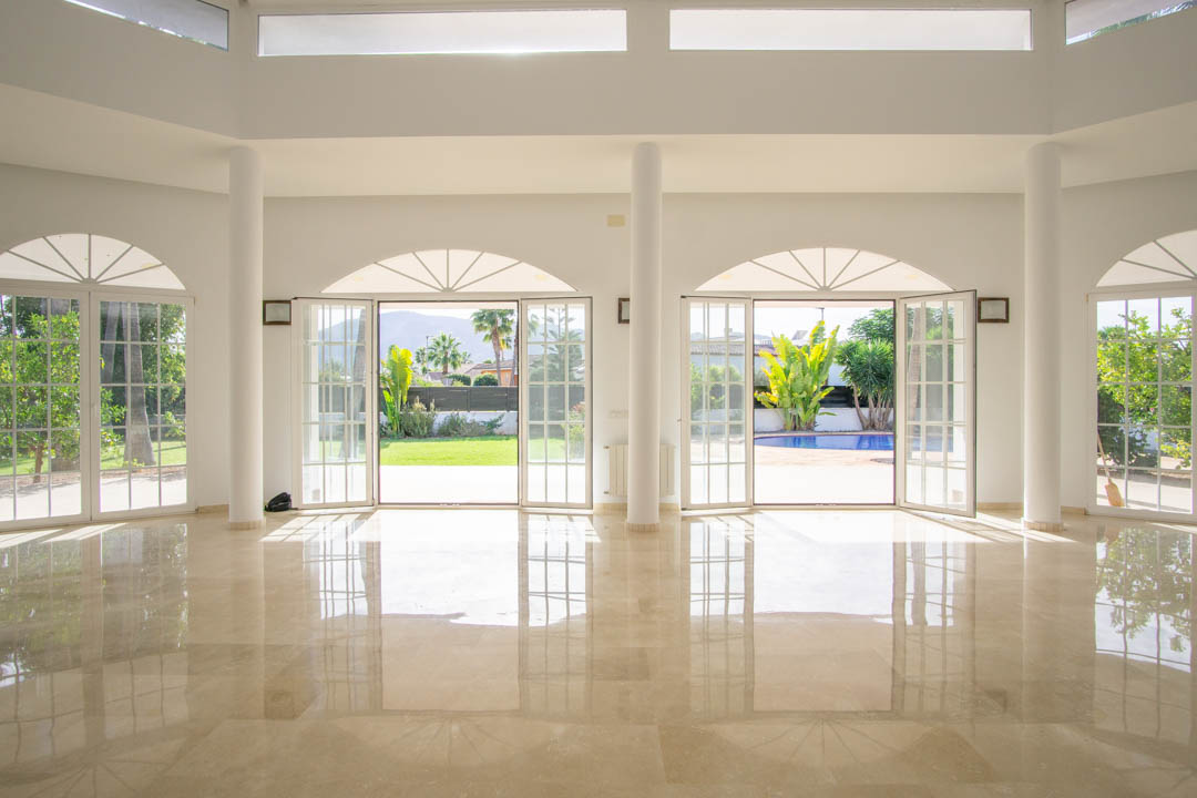 Amplio salón de estilo colonial con suelos de mármol pulido, columnas blancas y grandes ventanales de arco que dan acceso directo al jardín y la piscina.
