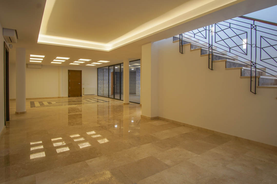 Amplio salón en planta sótano con suelos de mármol pulido, iluminación LED integrada y acceso a zona de bienestar acristalada junto a la escalera.