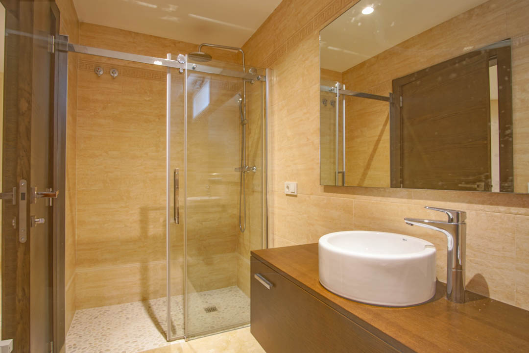 Baño moderno con ducha de mampara de cristal, suelo de cantos rodados, lavabo circular sobre encimera de madera y paredes alicatadas en tonos crema.