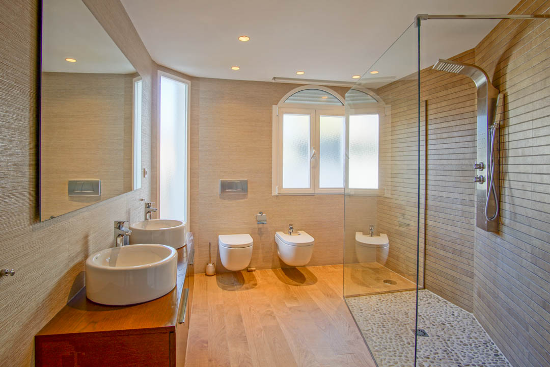 Baño moderno con lavabo doble sobre mueble de madera, ducha italiana con suelo de cantos rodados y sanitarios suspendidos bajo iluminación empotrada.
