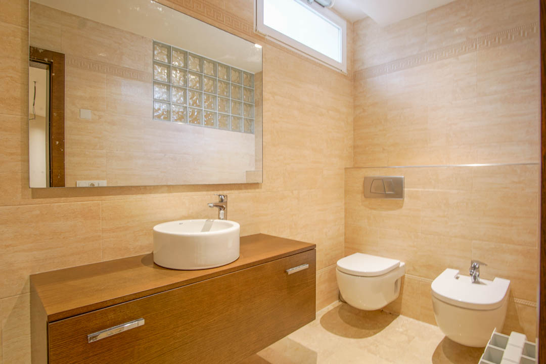 Baño revestido en tonos crema con mueble suspendido de madera, lavabo de sobremesa, sanitarios flotantes y espejo que refleja luz natural de bloques de vidrio.