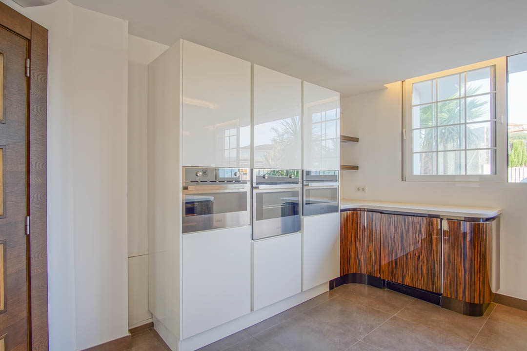 Cocina moderna con armarios blancos de alto brillo, hornos integrados de acero inoxidable y muebles bajos curvos con un distintivo acabado en madera veteada.