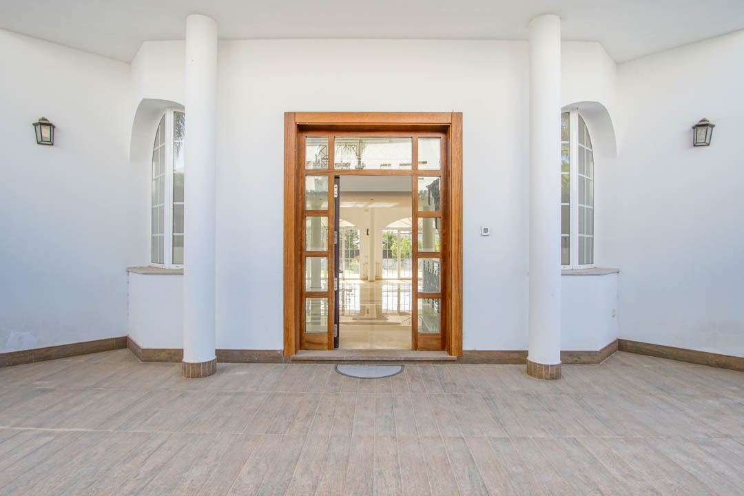 Entrada principal simétrica con puerta de madera y cristal, flanqueada por columnas blancas y ventanas de arco bajo un porche techado.