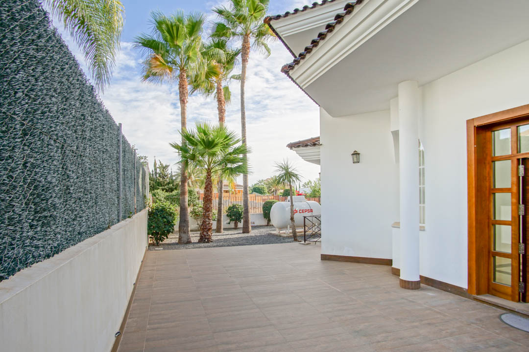 Exterior de la villa con porche de baldosas cerámicas, fachada blanca con columna circular y jardín lateral con palmeras altas junto al cerramiento.