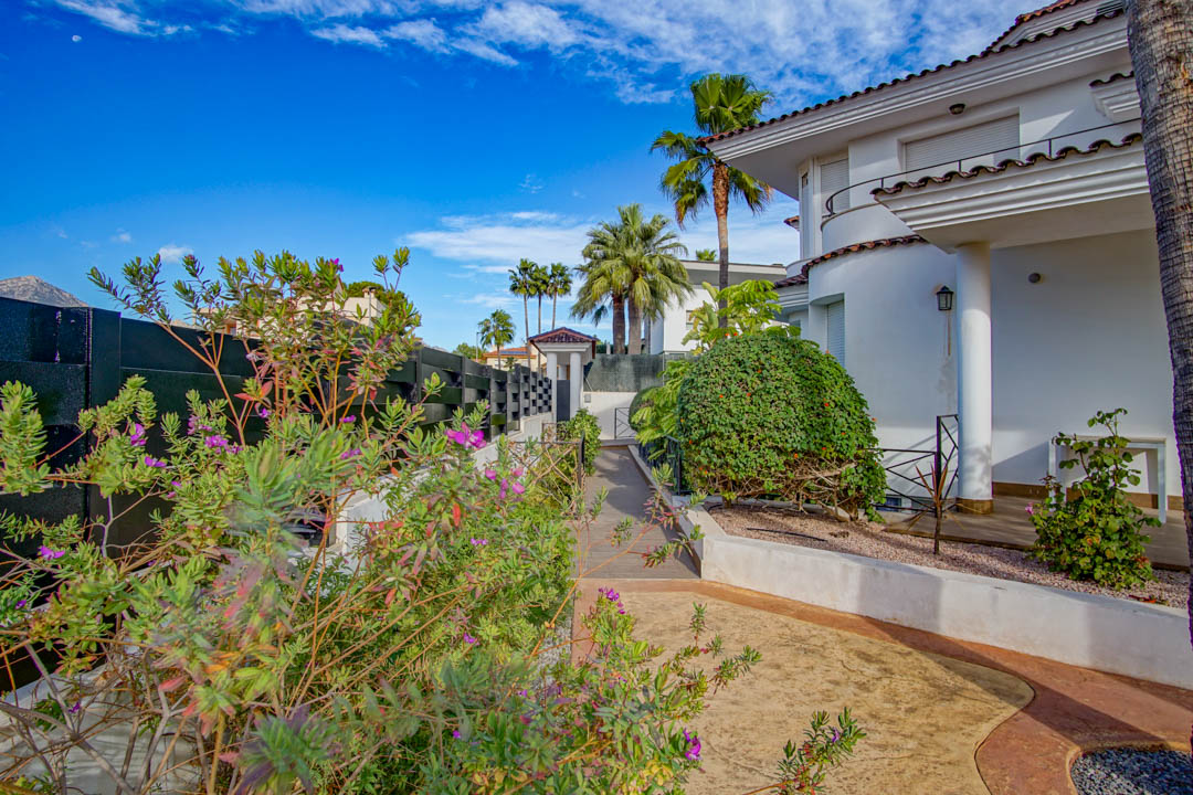 Fachada blanca de estilo colonial con columnas y porche, integrada en un jardín maduro con palmeras, arbustos florales y senderos pavimentados bajo un cielo despejado.