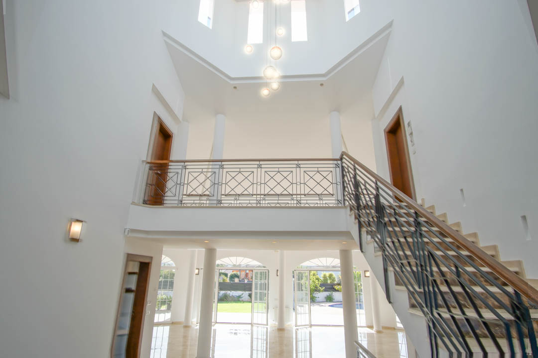 Hall de entrada de doble altura con escalera de mármol, barandilla de hierro forjado y una galería superior iluminada por ventanales altos y una lámpara colgante moderna.