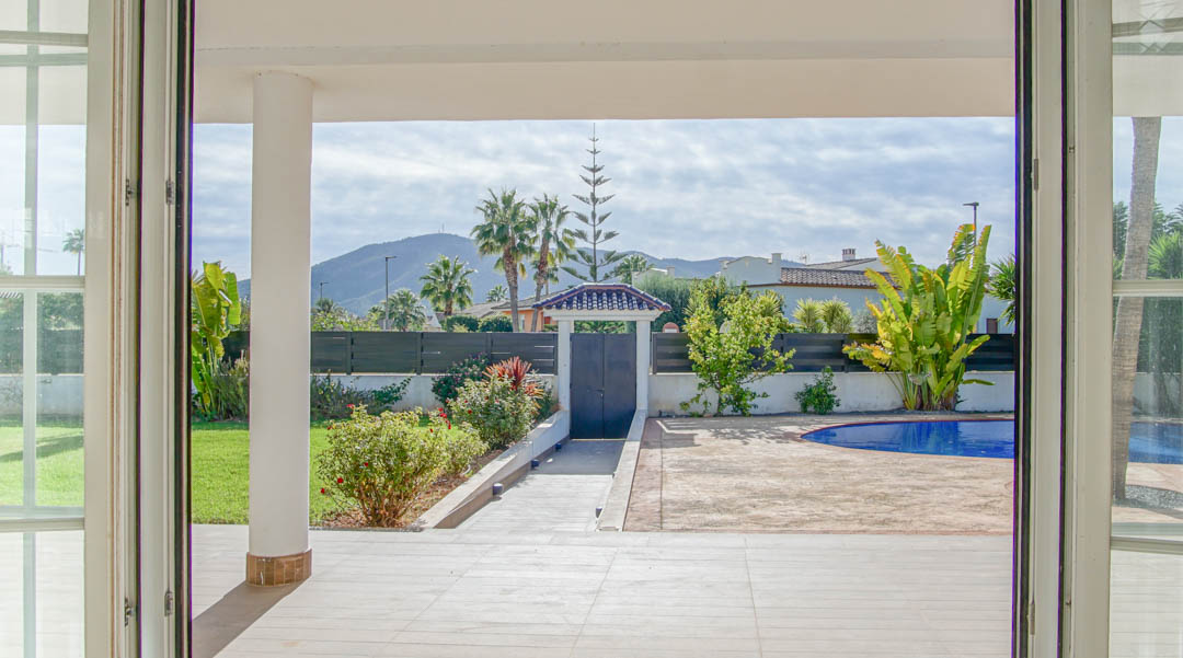 Vista desde el salón hacia el porche cubierto y el jardín, destacando la piscina azul y las vistas a la montaña a través de amplios ventanales blancos.