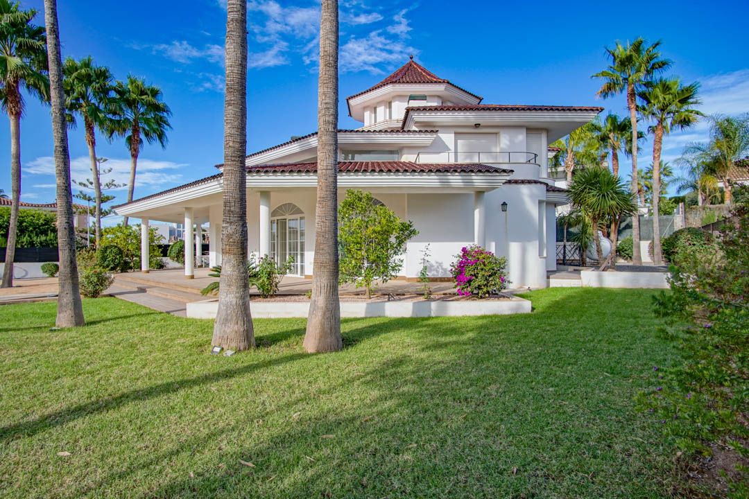 Vista exterior del chalet colonial con fachada blanca, jardín de césped cuidado y palmeras altas. Destaca el porche cubierto y los techos de teja tradicional.