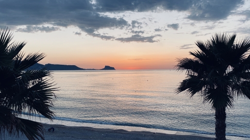 Vista panorámica del mar Mediterráneo al atardecer, enmarcada por palmeras desde la propiedad, con la silueta de la costa en el horizonte.