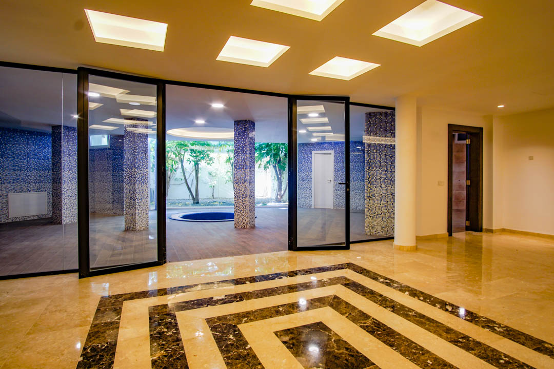 Zona de bienestar en el sótano con suelos de mármol decorados, columnas de mosaico azul y vistas a un patio inglés con árboles y jacuzzi integrado.