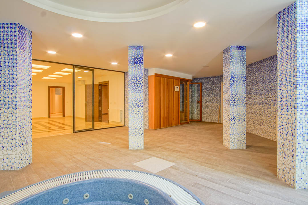 Zona de spa con jacuzzi circular, sauna de madera y columnas decoradas con mosaico azul. Incluye iluminación empotrada y acceso acristalado a otras estancias del sótano.