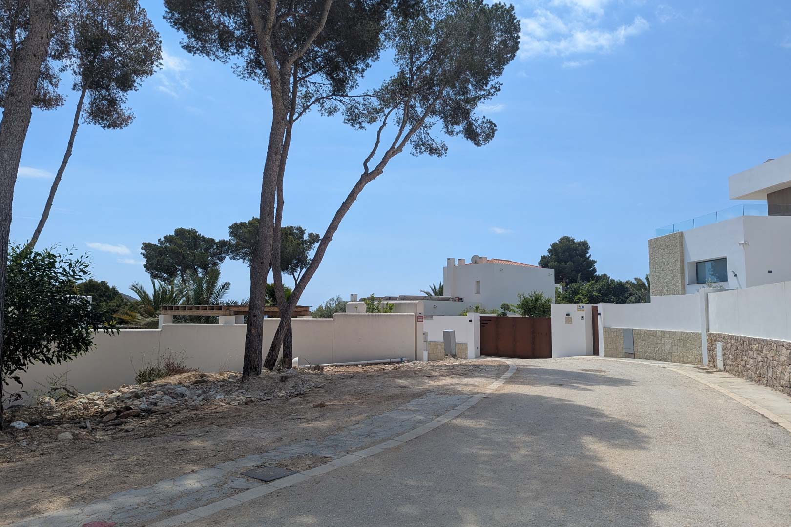 Vista de la calle de acceso en una urbanización tranquila de Moraira, rodeada de pinos maduros y muros perimetrales blancos de estilo mediterráneo.