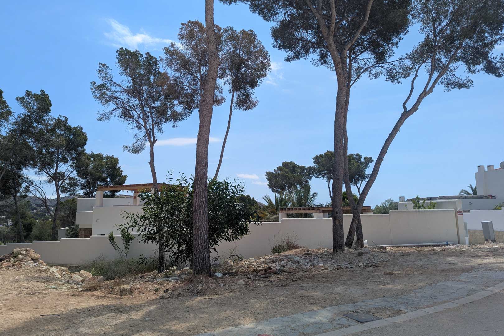 Vista de la parcela con pinos mediterráneos y muros perimetrales, donde se ubicará la villa de estilo ibicenco en un entorno residencial consolidado.