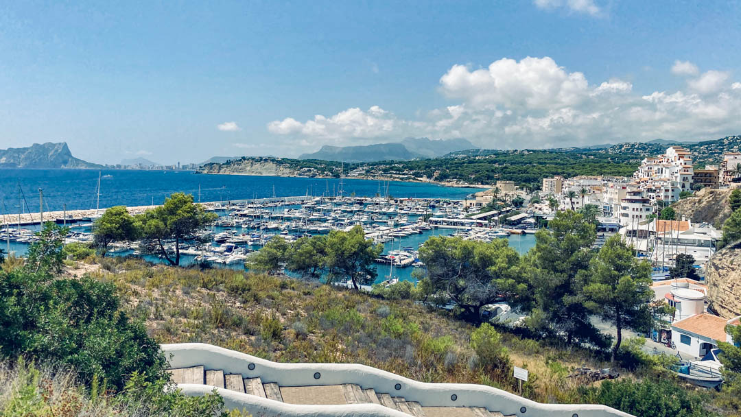 Vista panorámica del puerto deportivo de Moraira y el Peñón de Ifach desde una escalinata blanca de estilo mediterráneo rodeada de vegetación autóctona.