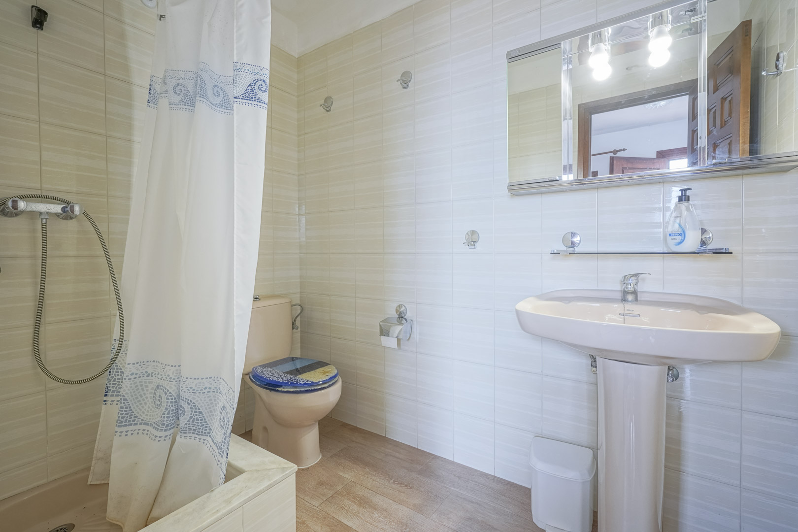 Cuarto de baño con azulejos claros, lavabo de pedestal rosa y cortina de ducha con motivos azules.