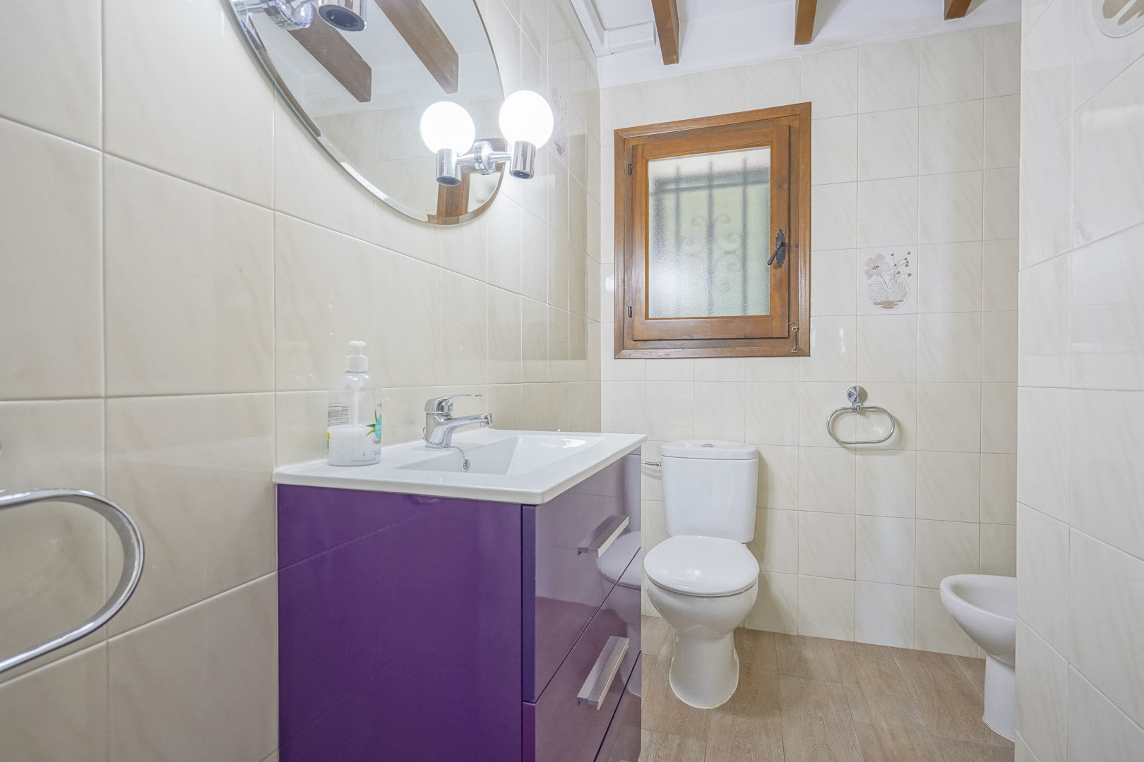 Detalle de un baño con azulejos color crema, lavabo morado y ventana con marco de madera.