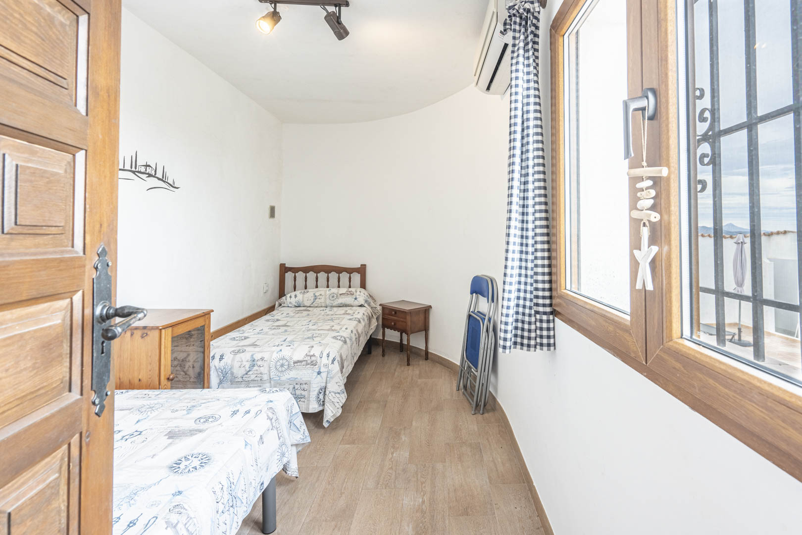 Dormitorio luminoso con dos camas individuales, decoración marinera y vistas exteriores desde la ventana enmarcada en madera.