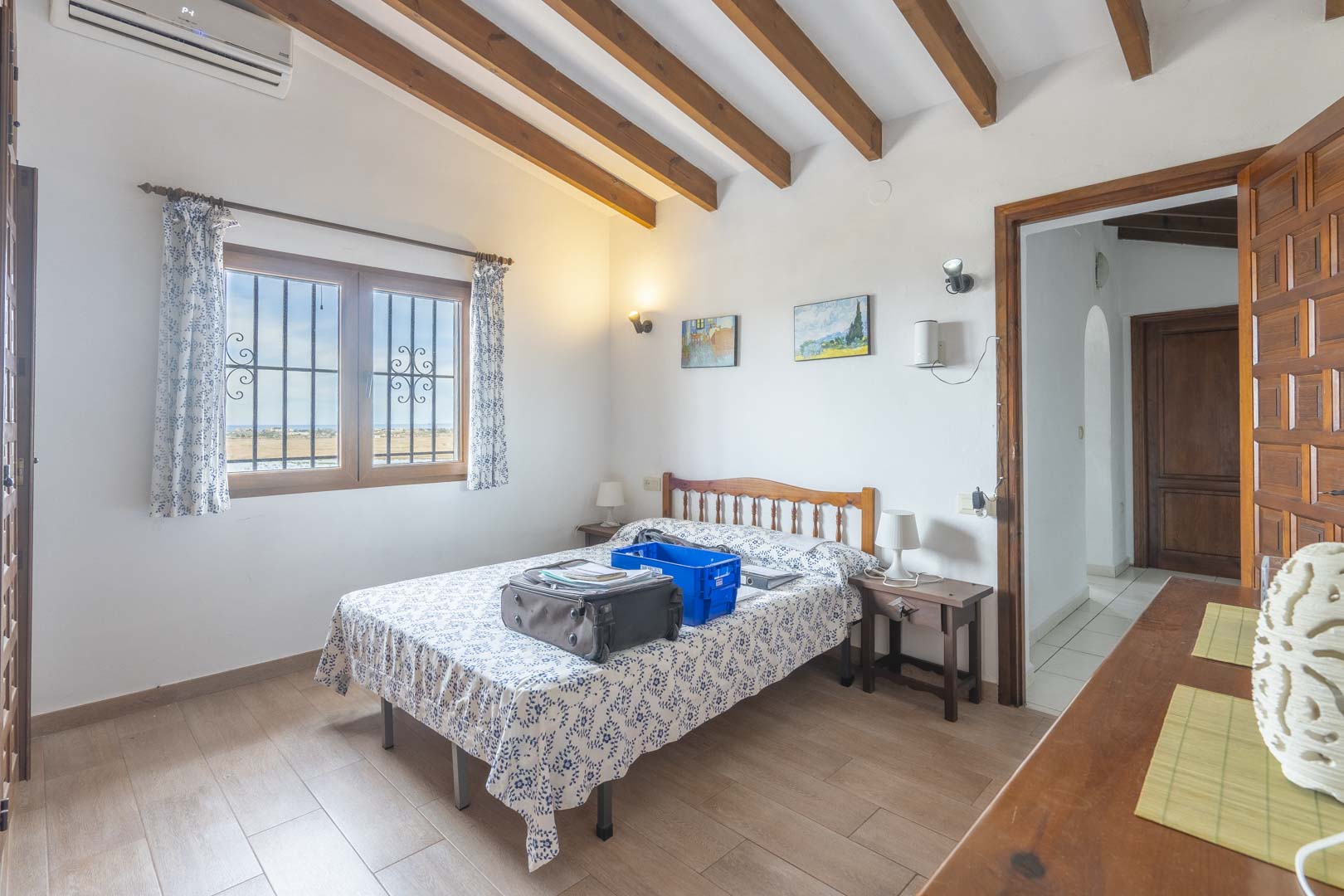 Dormitorio luminoso con vigas de madera, cama doble con ropa de cama estampada y vistas al exterior.