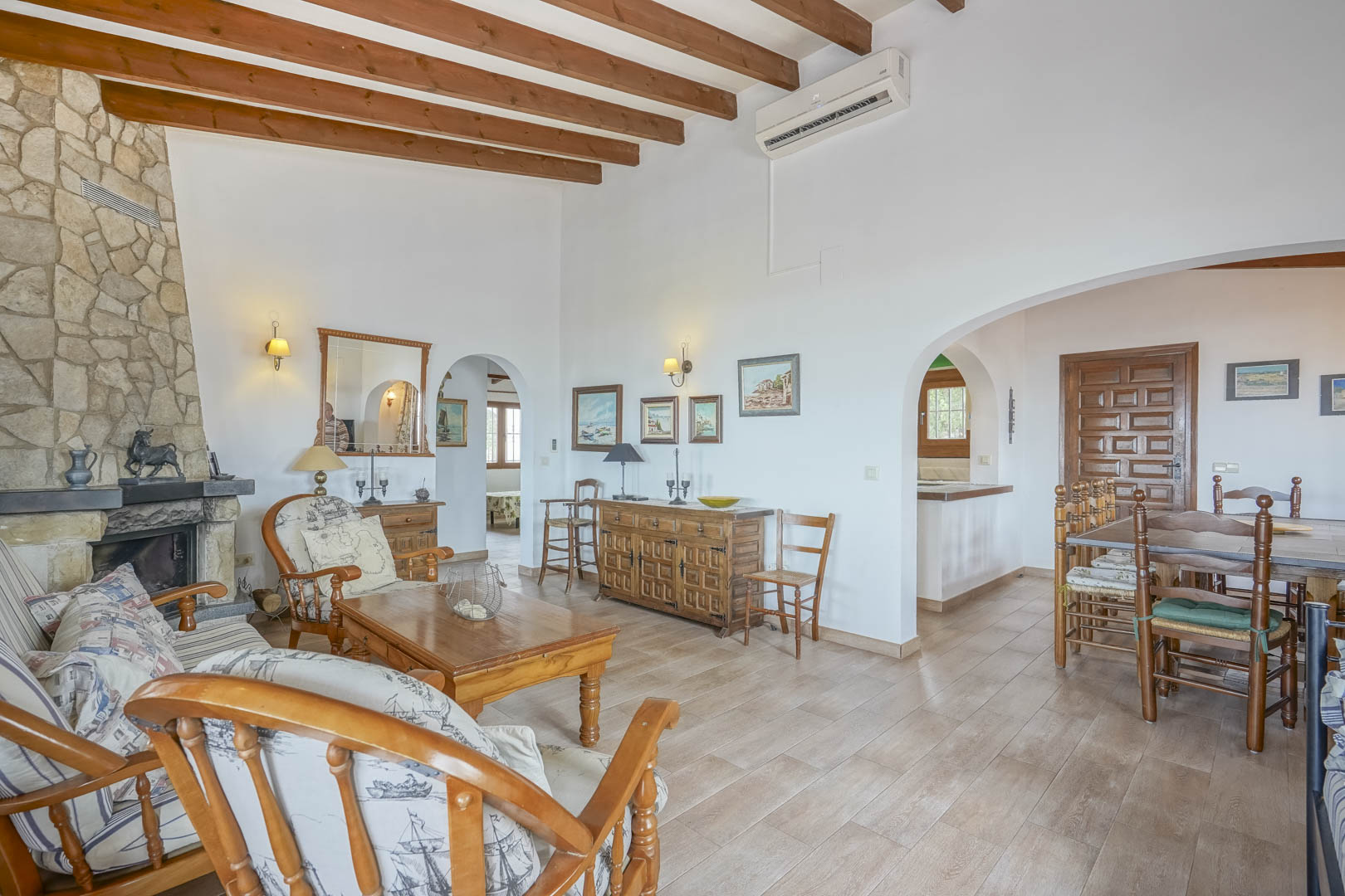 Salón con chimenea de piedra, vigas de madera vistas y mobiliario rústico. Espacios conectados por arcos.