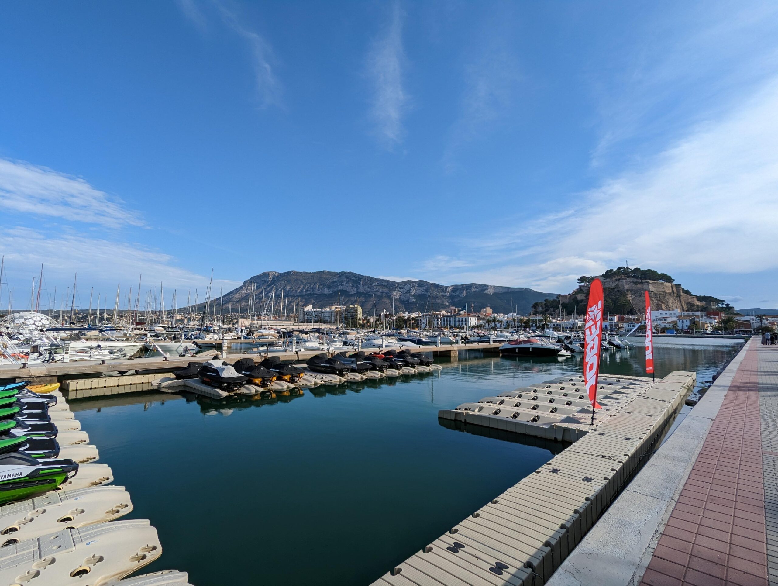 Vista del puerto deportivo con motos acuáticas y yates, con una montaña al fondo.