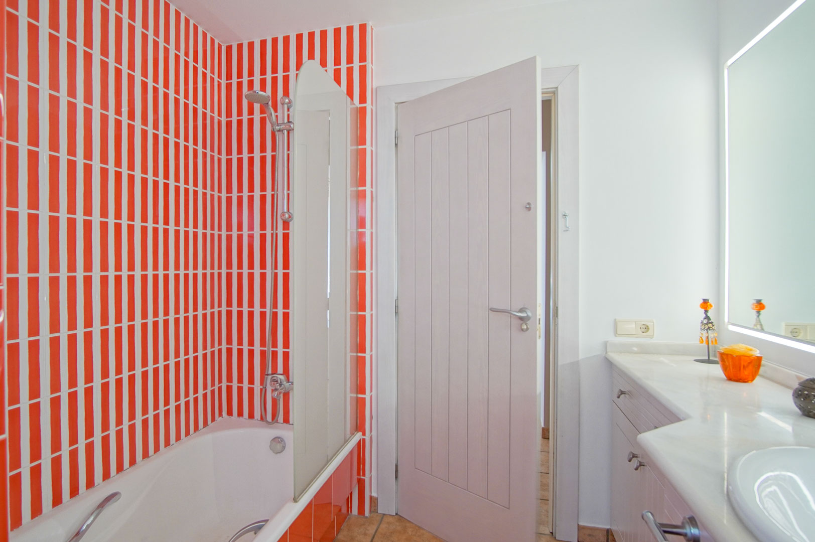 Baño con bañera protegida por mampara de cristal, resaltado por azulejos verticales en naranja y blanco, junto a un mueble con encimera de mármol y espejo con iluminación LED.
