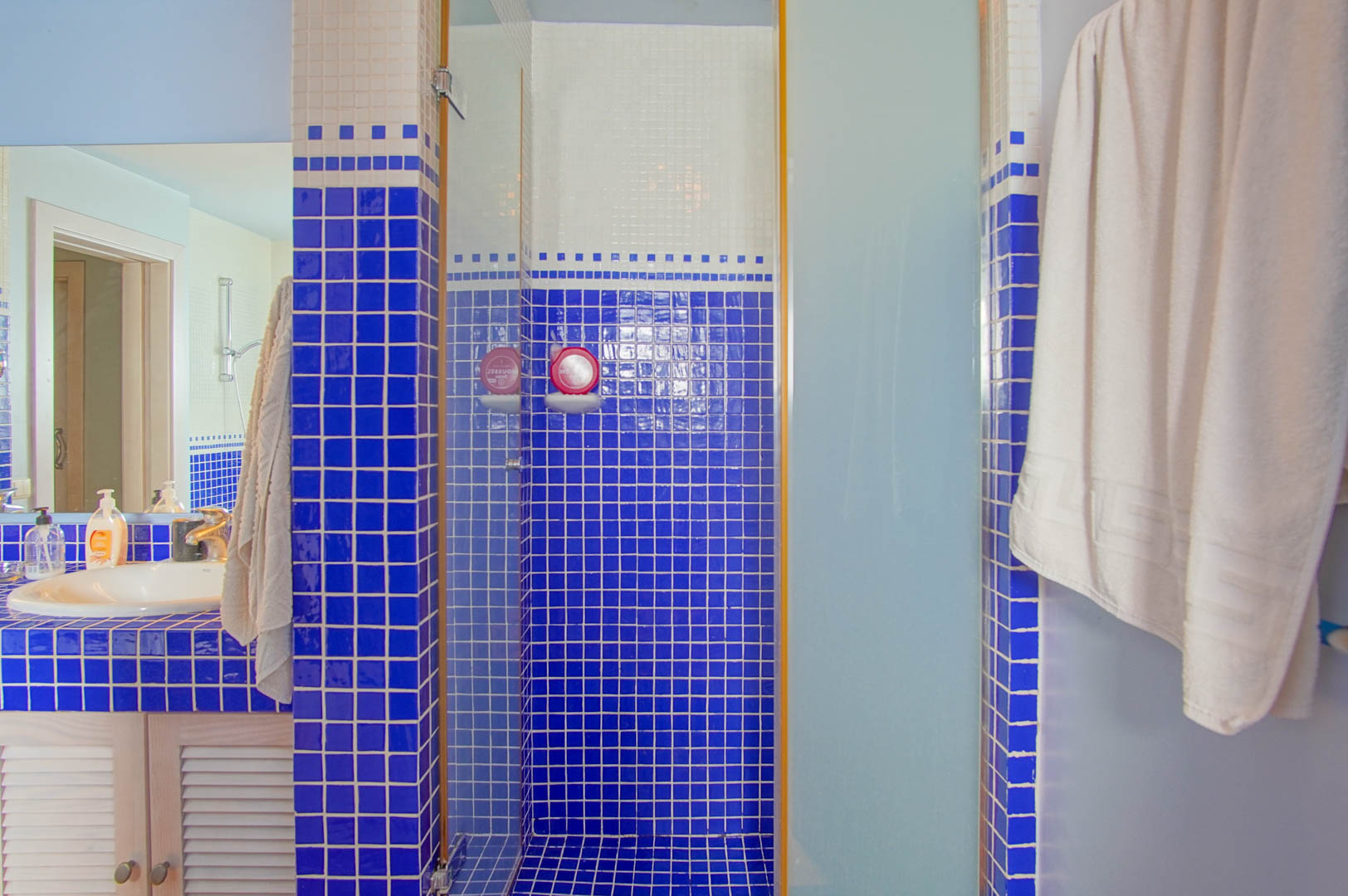 Baño con revestimiento de gresite azul y blanco, ducha de obra con mampara de cristal y lavabo integrado sobre mueble de madera con puertas de persiana.