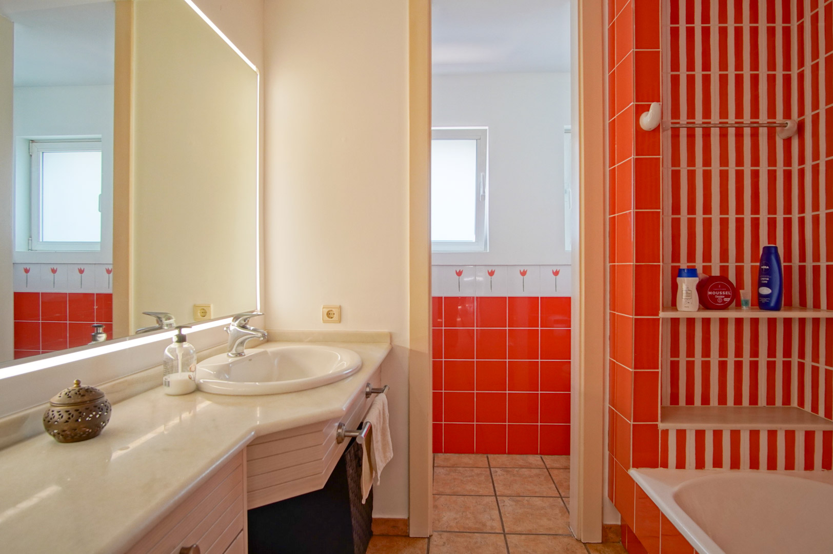 Baño funcional con encimera de mármol, espejo retroiluminado y azulejos en tonos naranja vibrante. Incluye zona de bañera con estantería integrada y entrada de luz natural.