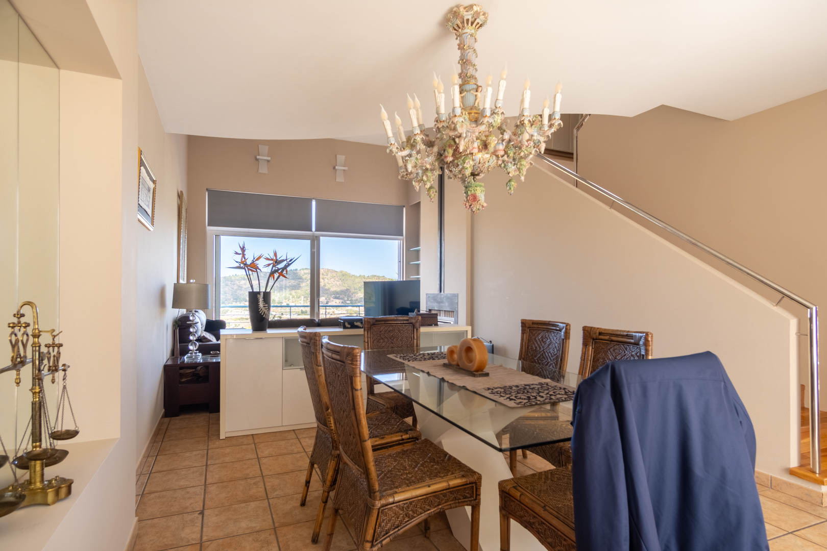 Comedor con mesa de cristal, sillas de mimbre y una lámpara de araña ornamental. Al fondo, ventanal con vistas a la montaña y escalera con barandilla metálica.
