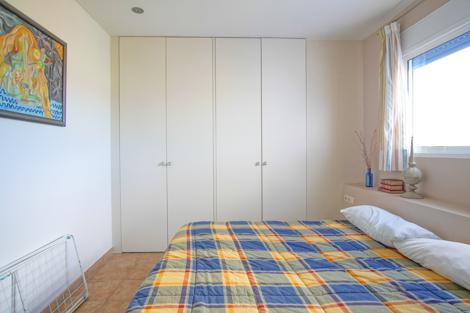 Dormitorio luminoso con amplios armarios empotrados blancos, suelos de gres rústico y una ventana lateral que permite la entrada de abundante luz natural.