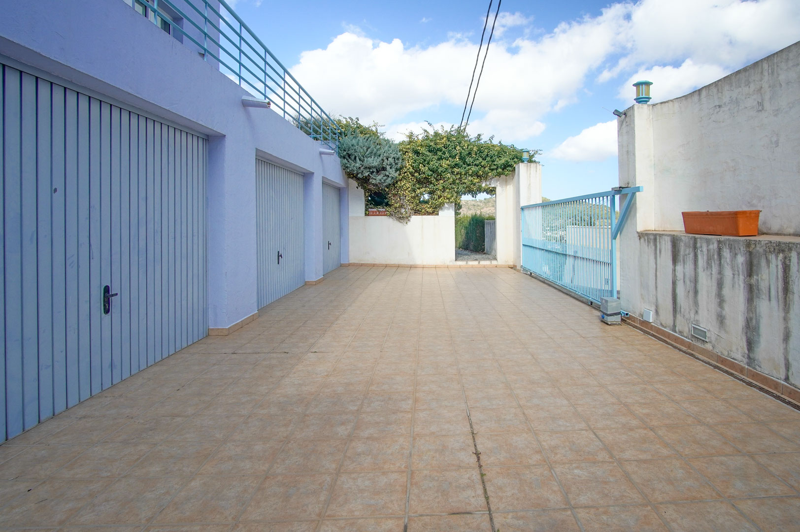 Entrada inferior con tres garajes privados de puertas azules, amplio acceso pavimentado y puerta corredera metálica, situada bajo la terraza superior de la villa.