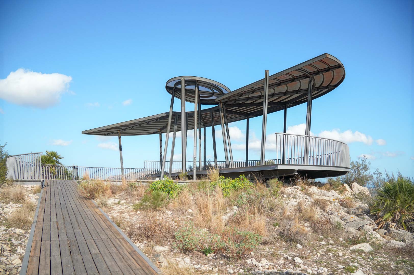 Mirador de diseño moderno con estructura metálica y cubierta curva, accesible mediante una pasarela de madera integrada en el entorno natural de la propiedad.