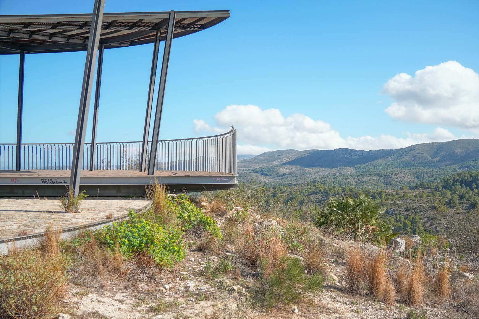 Mirador elevado con estructura metálica y barandilla de acero, ofreciendo vistas panorámicas despejadas hacia las montañas y el valle mediterráneo circundante.