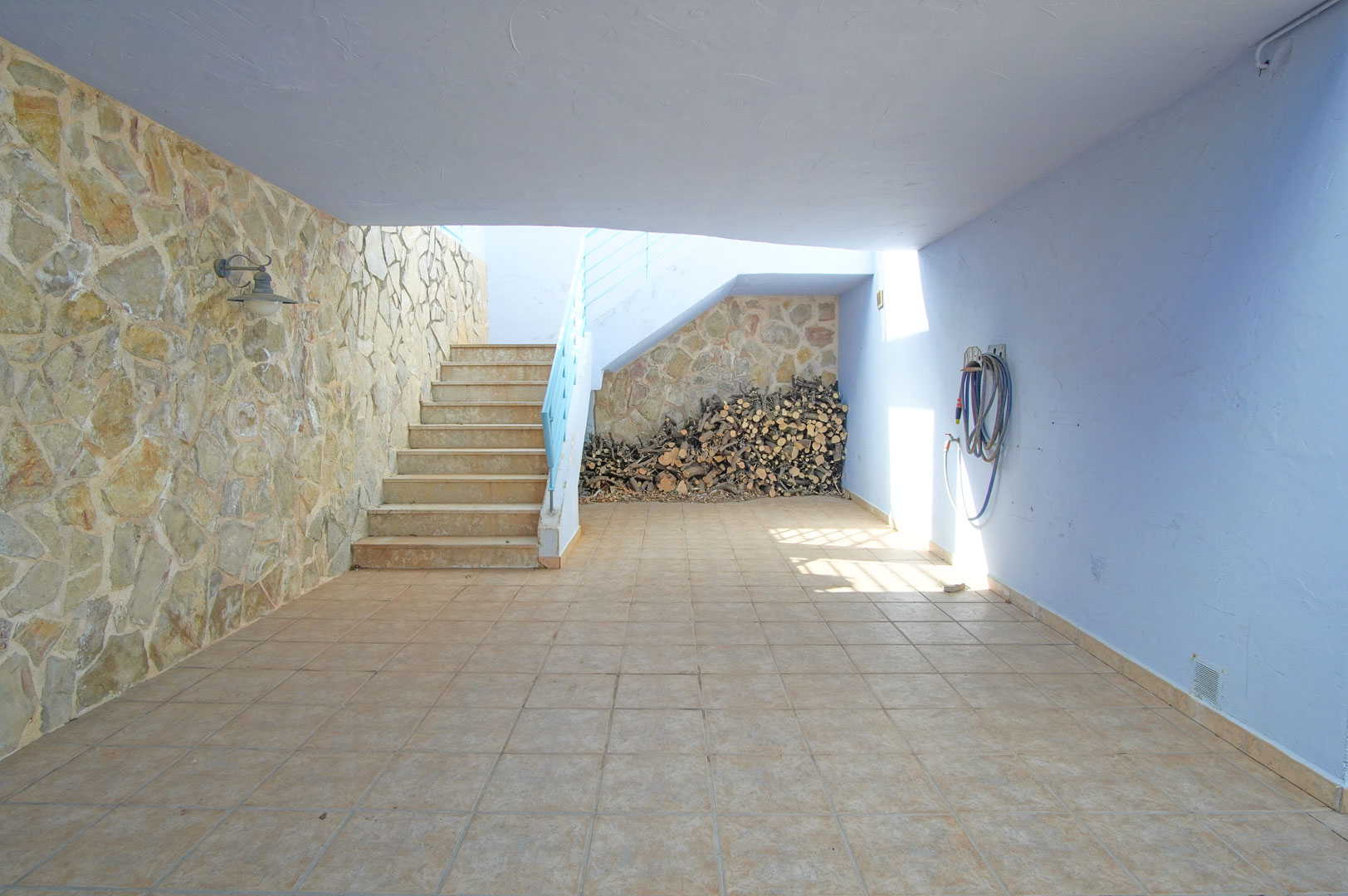 Patio cubierto con paredes de piedra natural, suelo cerámico y zona de almacenamiento de leña bajo la escalera exterior que da acceso a la planta superior.