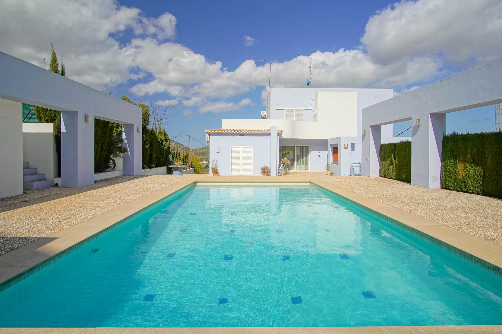 Piscina rectangular con mosaico turquesa, rodeada de una amplia terraza de grava y estructuras arquitectónicas modernas que enmarcan la fachada de la villa.